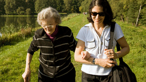 Eine alte Dame mit Brille und Stock ist bei einer jungen Frau mit Sonnenbrille und Handtasche eingehängt. Die beiden spazieren am Ufer eines Sees oder Flusses im Grünen.