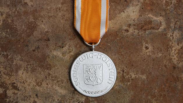 Silberne runde Medaille an einem orangefarbenem Band mit weißen Streifen.