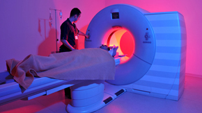 Ein Pfleger schiebt einen Patienten in ein Computertomographiegerät. Das Bild ist in leuchtenden Pink- und Violetttönen gehalten. Die Atmosphäre ist technisch-modern.