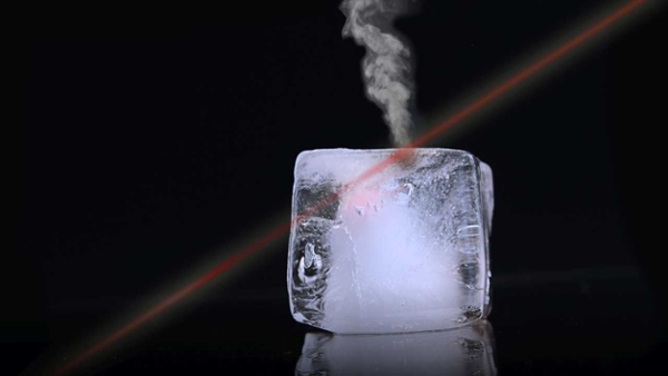 Rötlicher Laserstrahl trifft auf einen Eiswürfel, eine Rauchfahne steigt auf. Vor schwarzem Hintergrund.