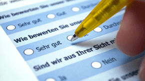 Mit einem gelben Kugelschreiber wird ein blauer Fragebogen ausgefüllt, auf dem mit schwarzer Schrift Fragen wie "Wie bewerten Sie unsere Qualität?" stehen.