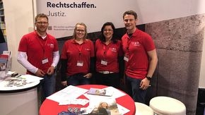 Zwei Männer und zwei Frauen stehen an einem Messestand. Im Vordergrund sind Tische mit Infomaterial.