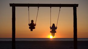 Zwei Kinder schaukeln bei Sonnenuntergang und schauen aufs Meer.