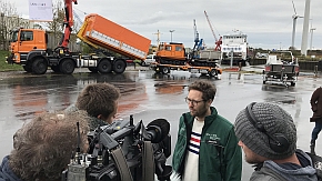 Umweltminister Jan Philipp Albrecht gibt ein Interview vor einer Fernsehkamera. Im Hintergrund ist ein Kran zu sehen, der ein Banner mit der Aufschrift "LKN.SH" hochhält.