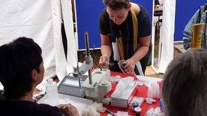 Eine Frau steht vor einem Tisch mit Laborgeräten und füllt mit einem Löffel dunkles Poulve aus einem Schraubglas in einem transparenten Plastikbecher.