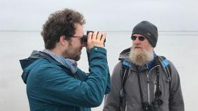 Zwei Männer stehen im Wattenmeer und unterhalten sich miteinander. Der eine schaut dabei durch ein Fernglas.