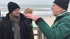 Zwei Männer unterhalten sich an einem Strand.