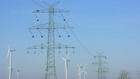 Große Strommasten vor blauen Himmer. Im Hintergrund sind Windräder zu sehen.