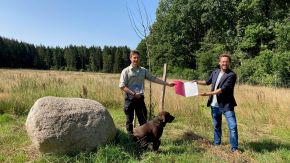 Zwei Männer stehen auf einer Wiese und halten gemeinsam eine Mappe mit einem Dokument in die Kamera. Im Vordergrun sitzt ein Hund.