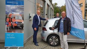 Staatssekretär Tobias Goldschmidt steht neben einem Elektrofahrzeug. Neben ihm steht ein Mann und steckt ein Ladekabel in die Ladebuchse des Autos.