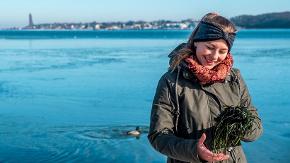 Eine Frau steht an einem Kieler Strand und hat angespültes Seegras in den Händen.