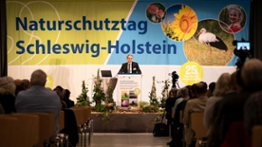 Ministerpräsident Daniel Günther steht auf einer Bühne und hält eine Rede vor mehreren Menschen. Im Hintergrund ist ein großes Banner mit der Aufschrift: Naturschutztag Schleswig-Holstein.