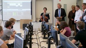 Eine Frau steht vor einem langen Tisch. An dem Tisch sitzen mehrere Jugendliche vor Computermonitoren.