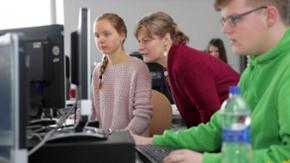 Im Computerraum einer Schule erklärt eine Lehrerin einer Schülerin etwas am PC, ein Schüler arbeitet selbständig daneben.