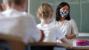 Eine Lehrerin steht vor der Tafel und trägt eine gemusterte Maske. 