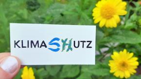 Das Bild zeigt einen Aufkleber mit der Aufschrift "KLIMASCHUTZ" vor gelben Blumen
