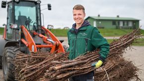 Ein junger Mann in grüner Arbeitskleidung trägt zwei große Bündel Äste unter beiden Armen. Im Hintergrund steht eine Baumaschine.