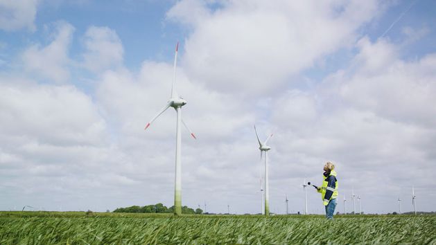 Eine Frau mit Schutzweste steht auf einem Feld und hält ein Gerät in die Luft. Im Hintergrund stehen Windräder.