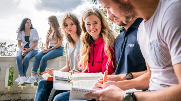 Mehrere junge Frauen und Männer sitzen gemeinsam im Freien und lernen für eine Prüfung. Auf ihren Knien liegen Bücher zum Steuerrecht. 