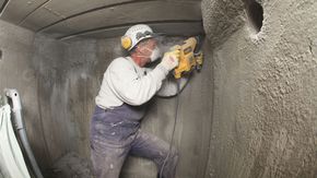 Ein Mann mit Helm, Ohrschützern und Mundschutz bearbeitet eine Betonwand mit einer Bohrmaschine.