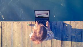 Eine Frau sitzt mit einem Laptop auf den Knien auf einem Bootssteg.