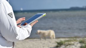 Eine Person hält am Strand ein Tablet in der Hand.