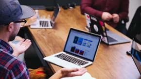 Ein Mann in Freizeitkleidung sitzt mit anderen Menschen an einem Konferenztisch und spricht über ein Projekt. Auf dem Tisch stehen mehrere aufgeklappte Laptops.