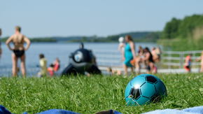 Ein Ball liegt im Gras. Im Hintergrund Menschen, die in einem See baden.