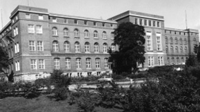 Schwarz-weiß-Aufnahme des Landtags vom Wasser aus gesehen. Der Glasbau für den Plenarsaal fehlt, vor dem Gebäude ist ein Parkplatz