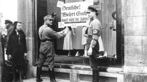 Historische Aufnahme zweier SA-Männer, die ein Plakat auf das Schaufenster eines jüdischen Bekleidungsgeschäfts kleben.