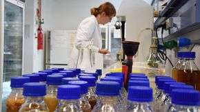 Im Vordergrund mit einer braunen Flüssigkeit gefüllte Flaschen, im Hintergrund steht eine Frau an einem Laborgerät