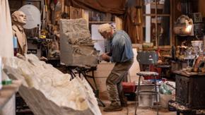 Ein älterer Mann steht in seinem Atelier und bearbeitet einen Steinblock. 