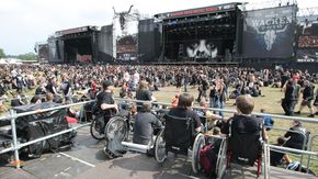 Drei Personen im Rollstuhl sitzen auf einer erhöhten Plattform, um die Bühne besser sehen zu können