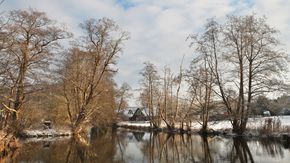 Ein Gewässer mit einem Haus im Hintergrund. Es liegt ein wenig Schnee.