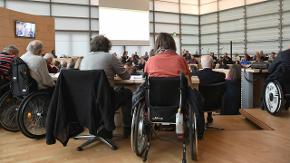 In einem großen Raum sitzen viele Menschen, einige davon im Rollstuhl.