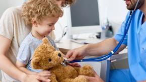 Ein Kind hält einen braunen Teddybär in den Händen. Ein Arzt hört das Herz vom Bären ab.