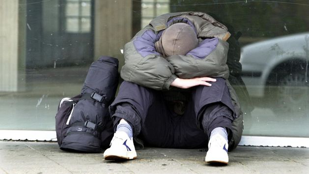 Ein junger Mann sitzt auf dem Boden und hat das Gesicht in seine Arme vergraben. Neben ihm steht ein Rucksack.