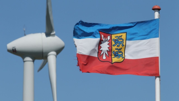 ein Windrad ist verschwommen neben der Flagge von Schleswig-Holstein zu sehen.