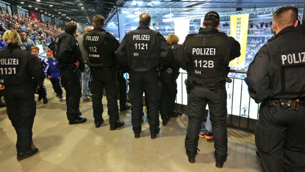 Sechs Polizeibeamte einer Einsatzhundertschaft stehen in einer Sporthalle und beobachten die Zuschauer.