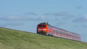 Roter Zug auf grasbewachsenem Damm vor blauem Himmel